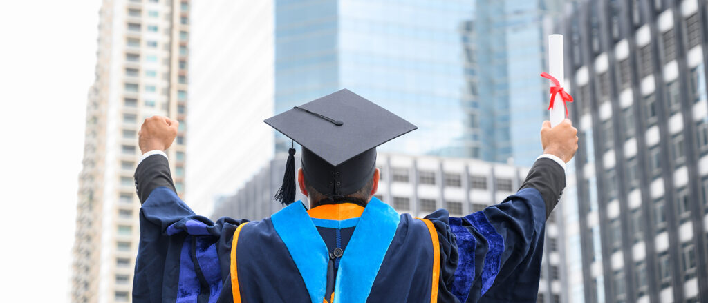 A man in a blue graduation cap and gown holds up a diploma triumphantly, his back to the camera. Several tall buildings fill up the background.