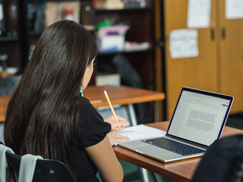 An Indigenous student writes in a notebook, a laptop open on her desk.