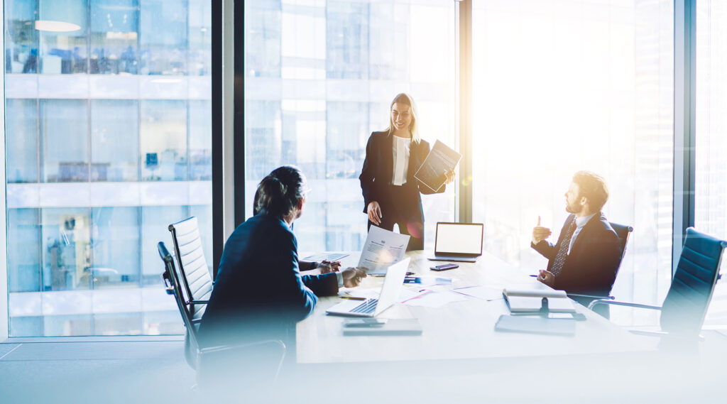 miling female executive manager talking to colleagues in boardroom in front of a wall of windows on a sunny day