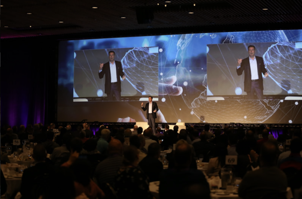 A man speaking on a stage, with two video screens behind him